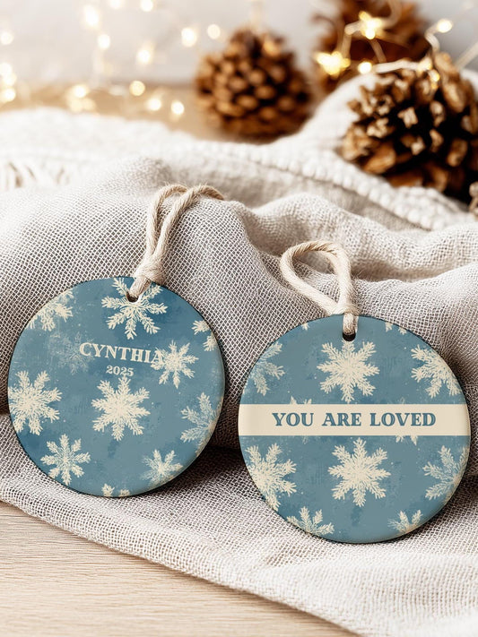 Vintage blue ceramic snowflake ornament with personalized name, displayed on soft beige linen with pinecones and warm fairy lights in the background.