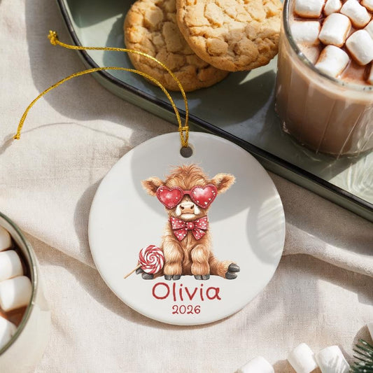 May include: A white ceramic ornament featuring a Highland cow illustration wearing heart-shaped sunglasses and a bow tie. The ornament includes the name "Olivia" and the year "2026" in red script, with a gold string for hanging.