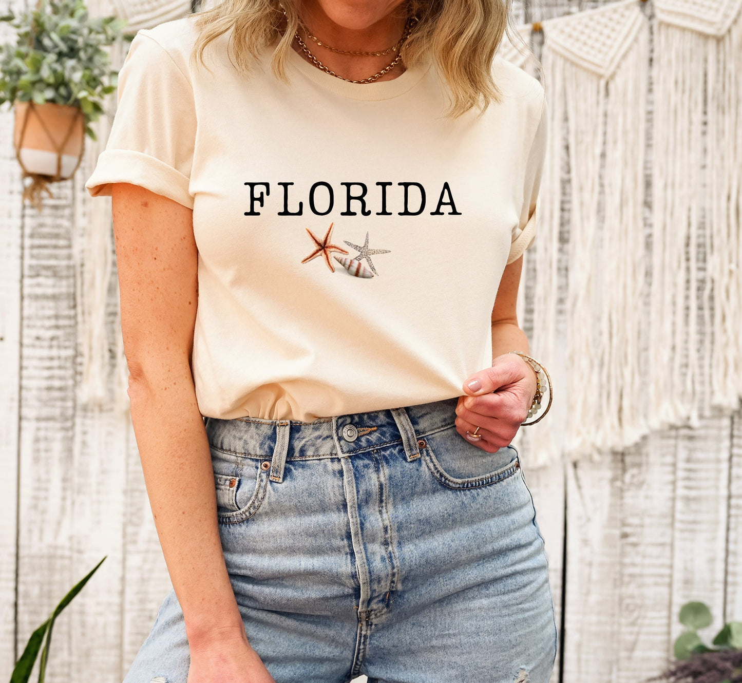 May include: A cream colored t-shirt with the word 'FLORIDA' printed in black text. The shirt has a small image of a seashell and a starfish printed below the text.