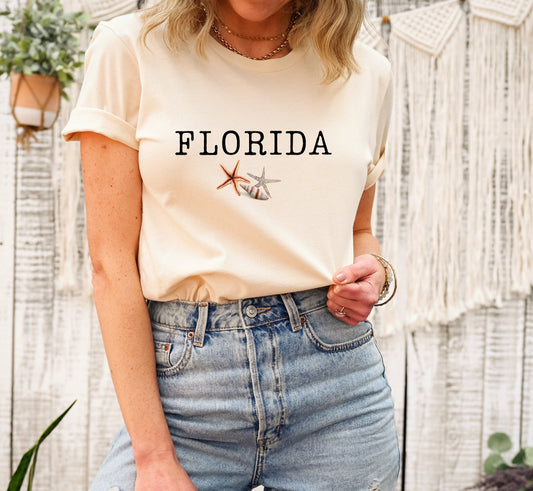 May include: A cream colored t-shirt with the word 'FLORIDA' printed in black text. The shirt has a small image of a seashell and a starfish printed below the text.