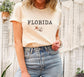 May include: A cream colored t-shirt with the word &#39;FLORIDA&#39; printed in black text. The shirt has a small image of a seashell and a starfish printed below the text.