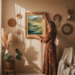 High resolution 2:3 ratio digital download poster is in a simple wood frame, The lady is fixing the frame on her beige color wall. The wall has other bohemian accessories such as greenery plants, macrame designs. Her flowery dress in terracota color and a sofa shawl is next to her. The theme is cozy, boho and lively.