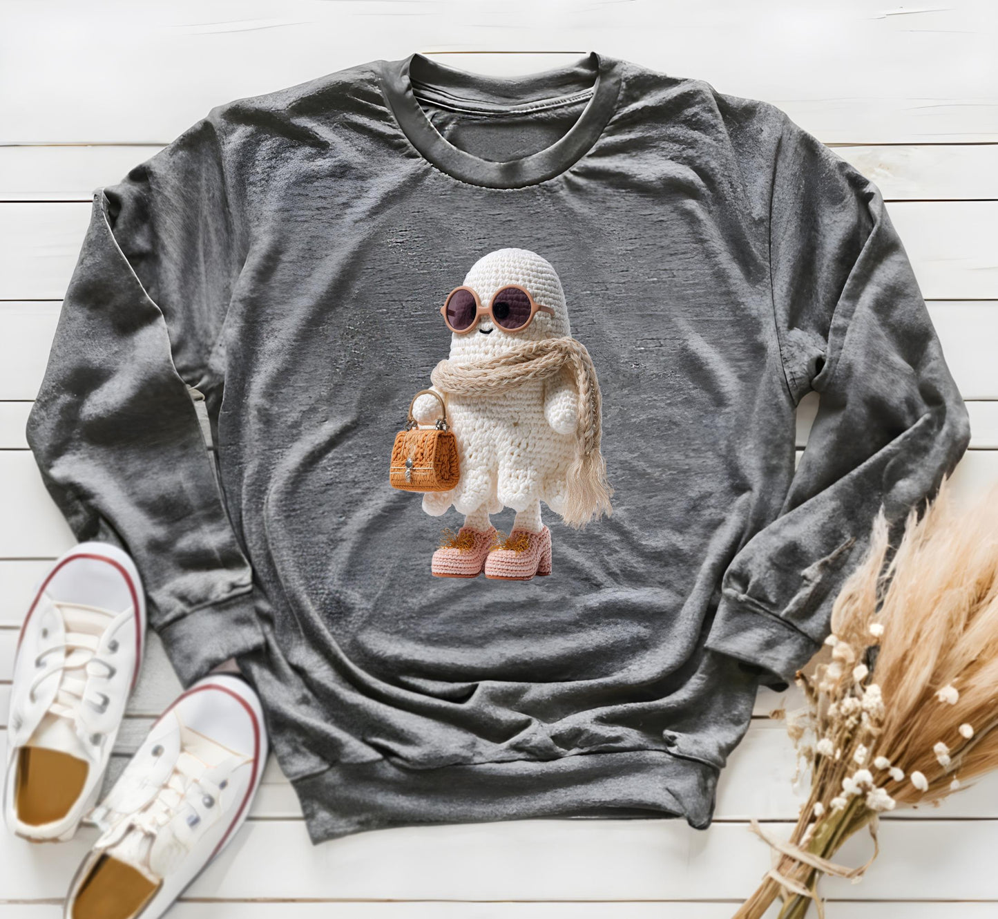 Coquette latch yarn ghost PNG is placed on a Gildan dark grey sweatshirt on a distressed surface. The mockup has white sport shoes and a dried bouquet on the right side. The Boojee ghost is white, blush pink and leather colors so it is noticeable.