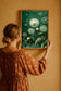 Flowery dressed bohemian woman with loose hair bun is holding the framed digital poster on a warm color wall. She is looking at the emerald green background dandelion wall art. There is a perfect macthing fall colors combination in the photo.