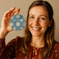 A woman with a big smile is holding a vintage blue and beige colored snowflake ornament with custom name and year.