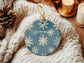 Ceramic round ornament in blue with snowflake pattern and the text “You Are Loved,” behind the ornament and styled on textured fabric with cozy holiday decor.