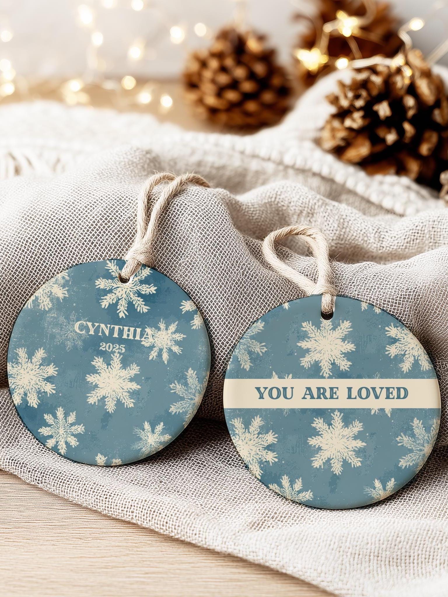 Vintage blue ceramic snowflake ornament with personalized name, displayed on soft beige linen with pinecones and warm fairy lights in the background.