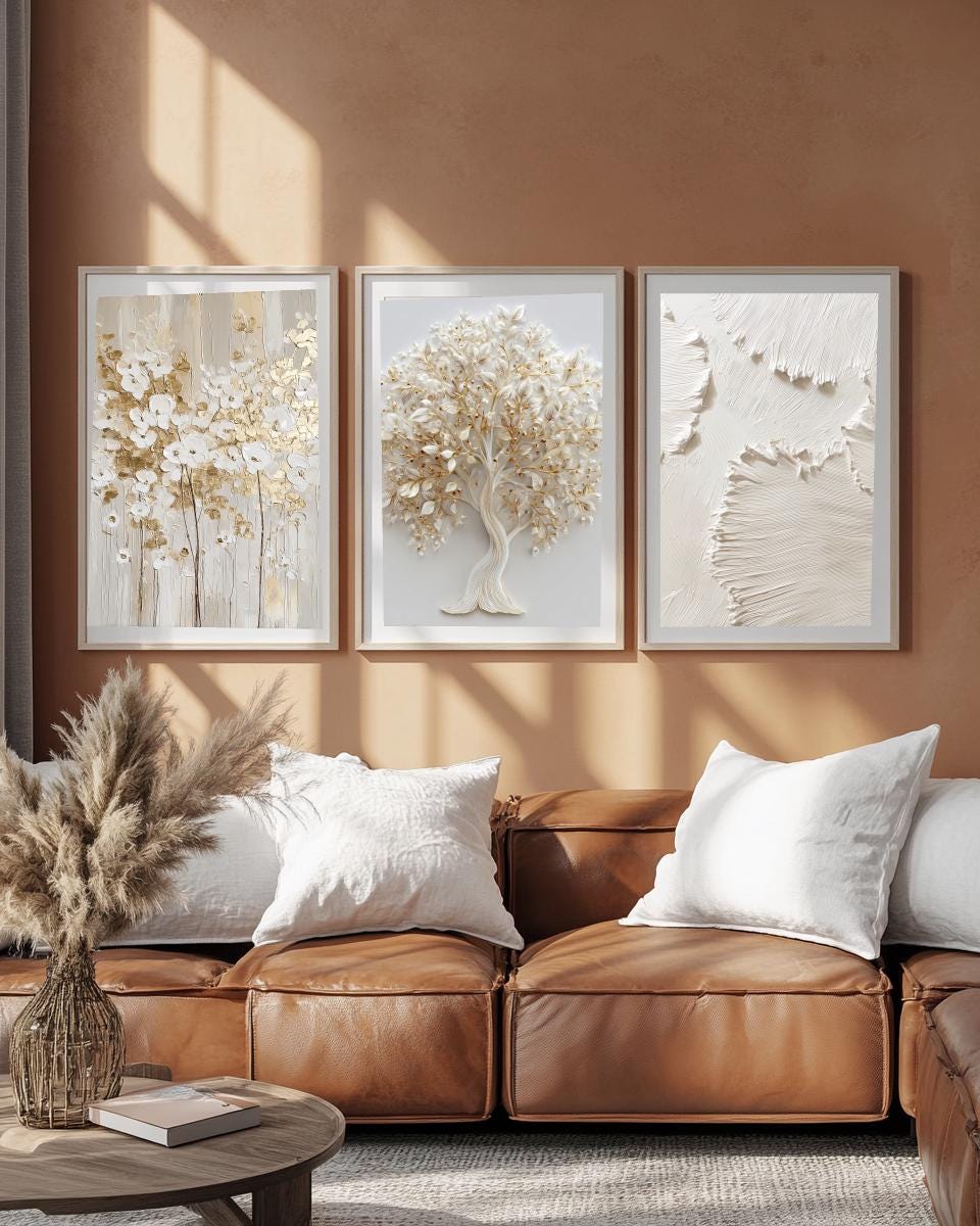 Textured 3 wall art design hanging on a beige wall. The room has leather loveseat with some daylights. One of the frame is textured minimal brushstrokes, The wall art in the middle is with white and gold color elegant tree, the wall art on the right side has golden white floral oil painting with golden foil effect.