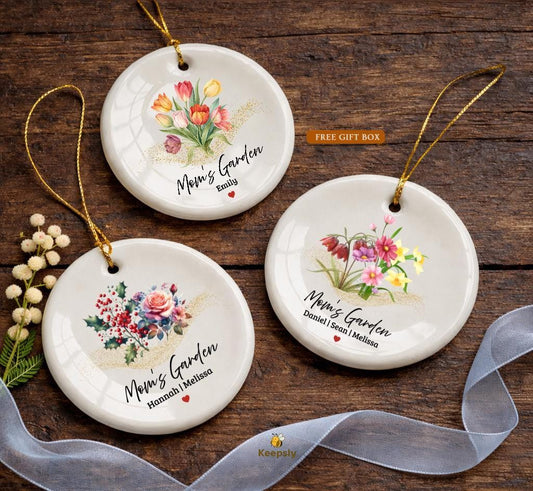 May include: Three white ceramic ornaments with floral designs and gold hanging cords. Each ornament features a different flower arrangement and the text "Mom's Garden" with a name below. The ornaments are on a wooden surface.