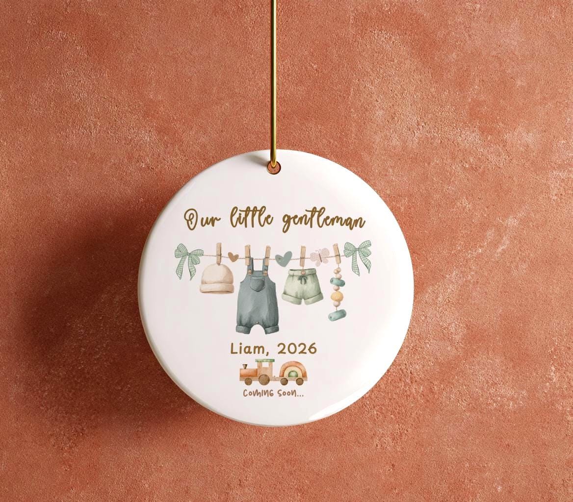May include: A round, white ceramic ornament with a gold string. The ornament features a watercolor illustration of baby clothes hanging on a clothesline, with the text "Our little gentleman, Liam, 2026, Coming Soon..."