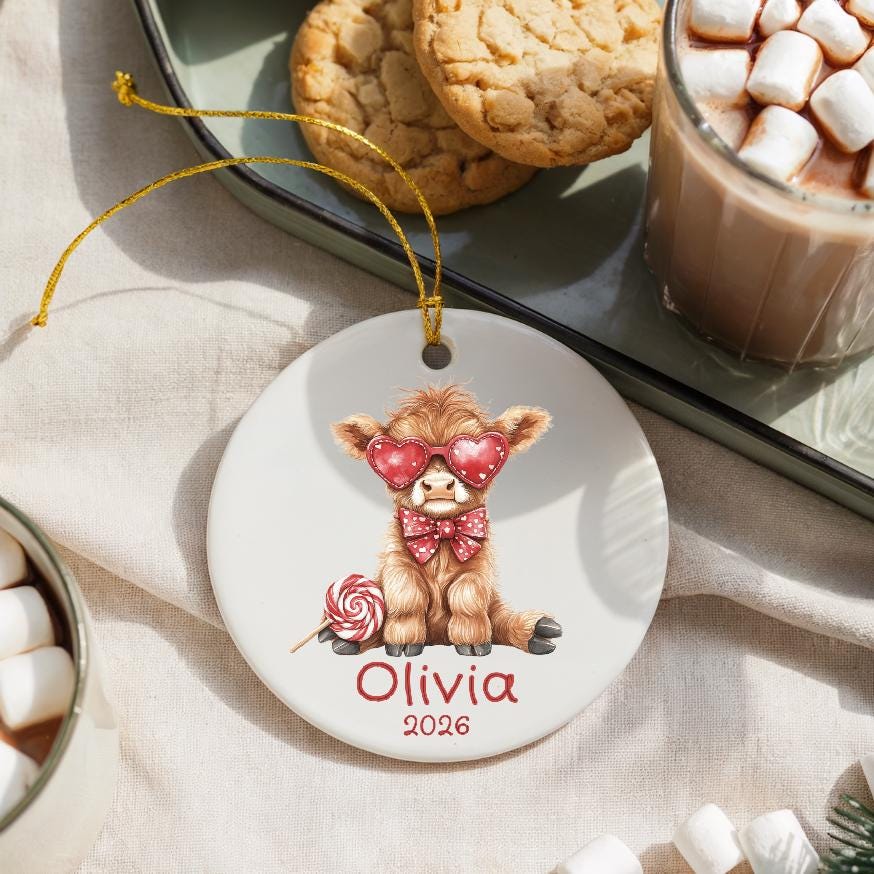 May include: A white ceramic ornament featuring a Highland cow illustration wearing heart-shaped sunglasses and a bow tie. The ornament includes the name "Olivia" and the year "2026" in red script, with a gold string for hanging.
