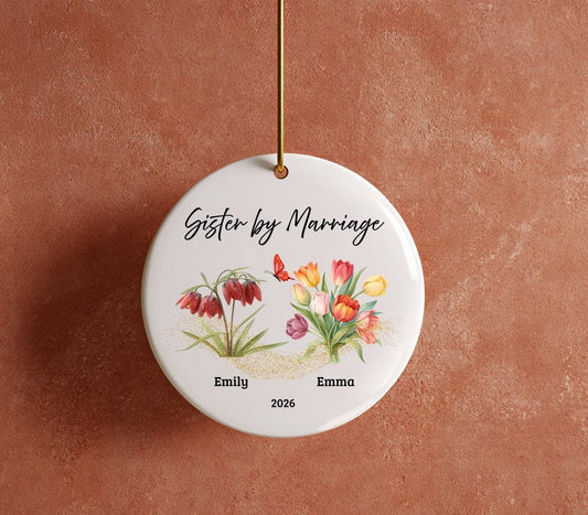 May include: A round, white ceramic ornament with the text "Sister by Marriage" in black script. The ornament features watercolor illustrations of flowers and a butterfly, with the names "Emily" and "Emma" and the year "2026" below.