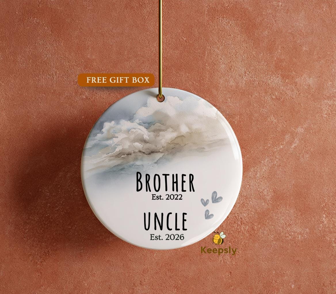 May include: A round, white ceramic ornament with a watercolor cloud design. The ornament reads "BROTHER Est. 2022" and "UNCLE Est. 2026" in black text. Small blue hearts and a gold string are also visible.