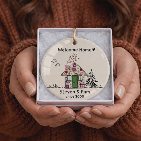 Close-up of hands holding a custom ceramic welcome home ornament in a gift box, featuring floral watercolor artwork, a green door, personalized family names, and the year.