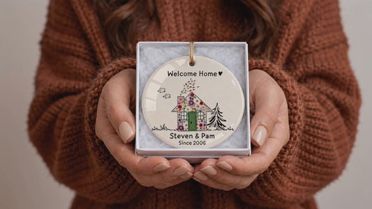 Close-up of hands holding a custom ceramic welcome home ornament in a gift box, featuring floral watercolor artwork, a green door, personalized family names, and the year.
