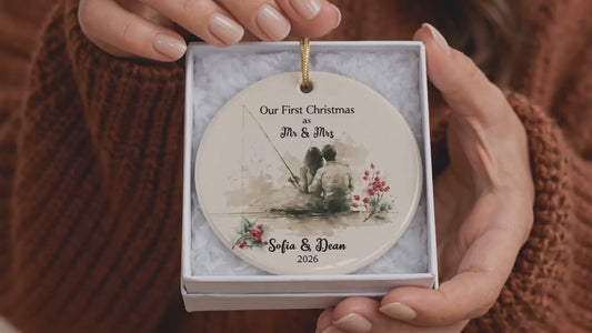 Close-up of hands holding a custom ceramic first Christmas married fishing ornament in a gift box, featuring a watercolor illustration of an outdoor couple fishing, personalized names, and wedding date